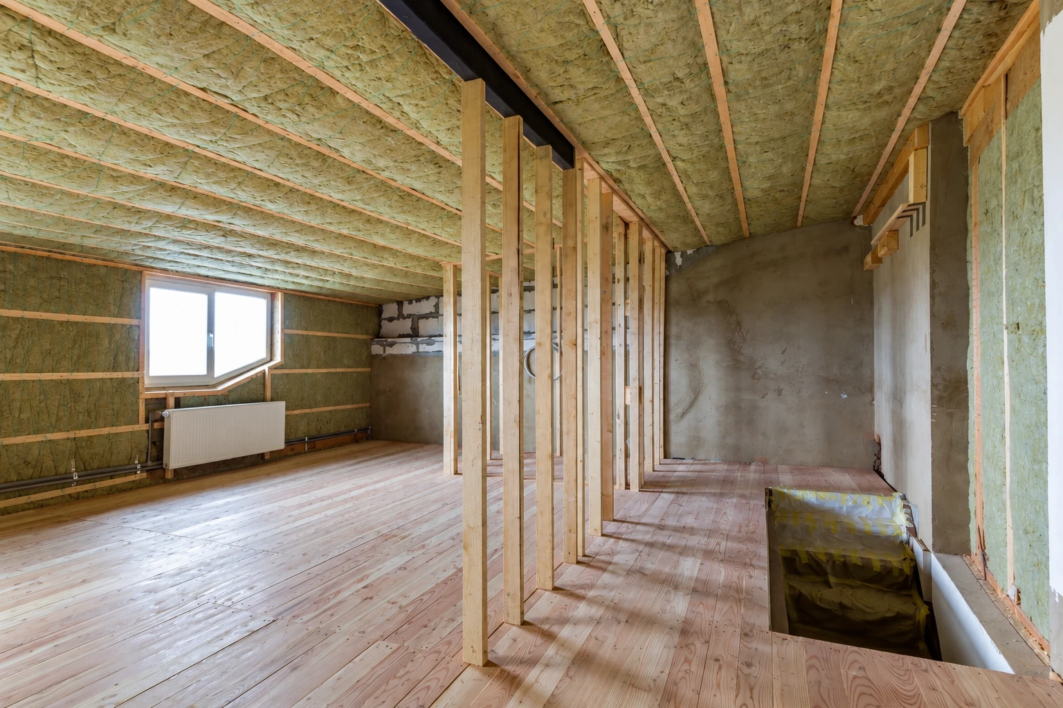 Attic with green insulation between rafters and wall studs, a window, and unfinished wooden flooring.