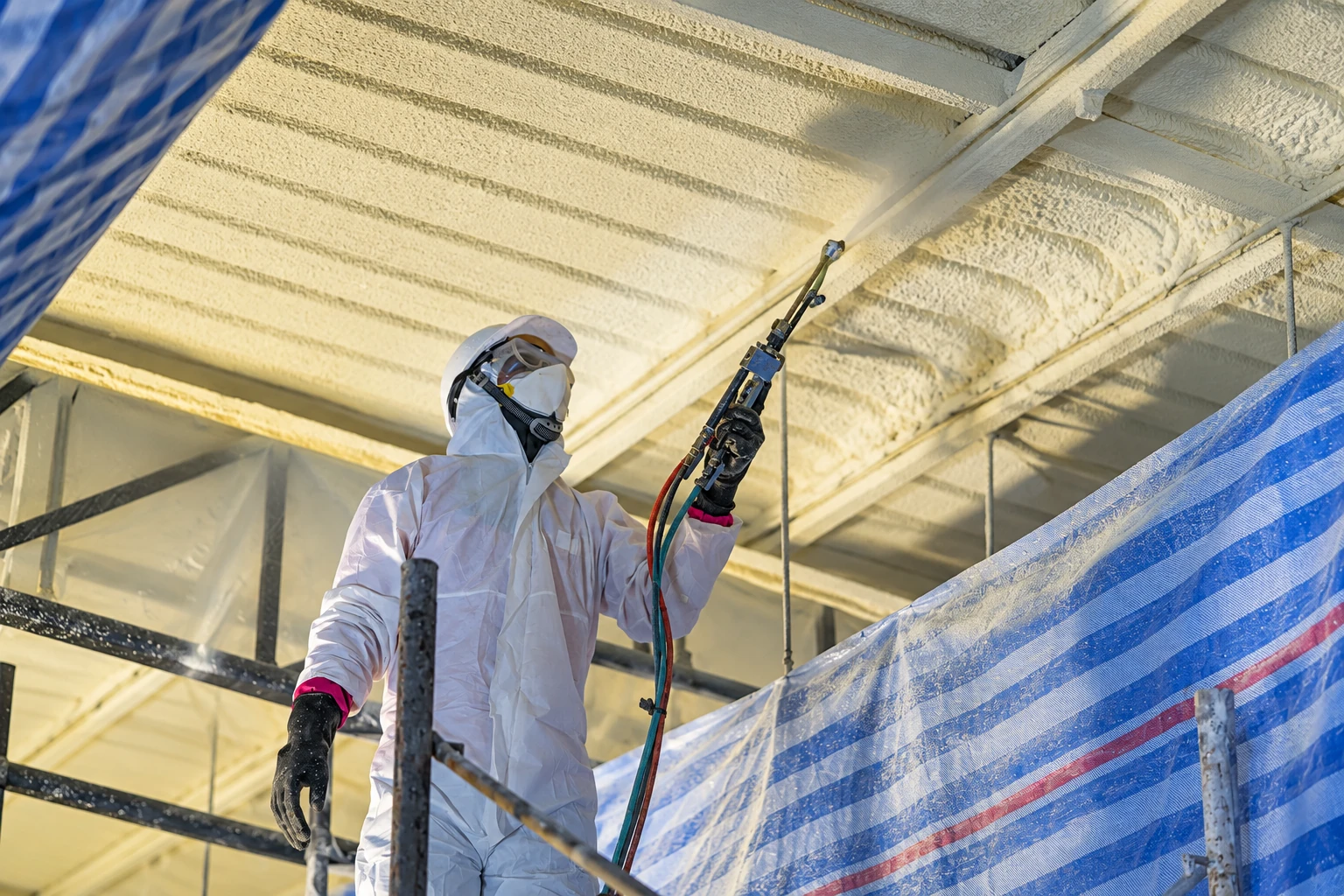 A worker in protective gear sprays closed-cell foam onto the ceiling of a construction site.