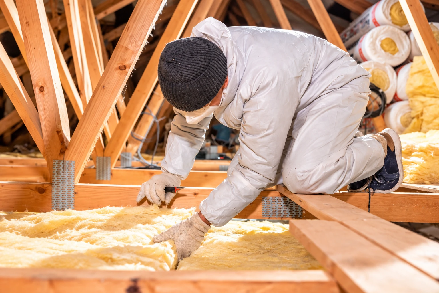Worker in protective clothing lays yellow fiberglass insulation between wooden attic joists.