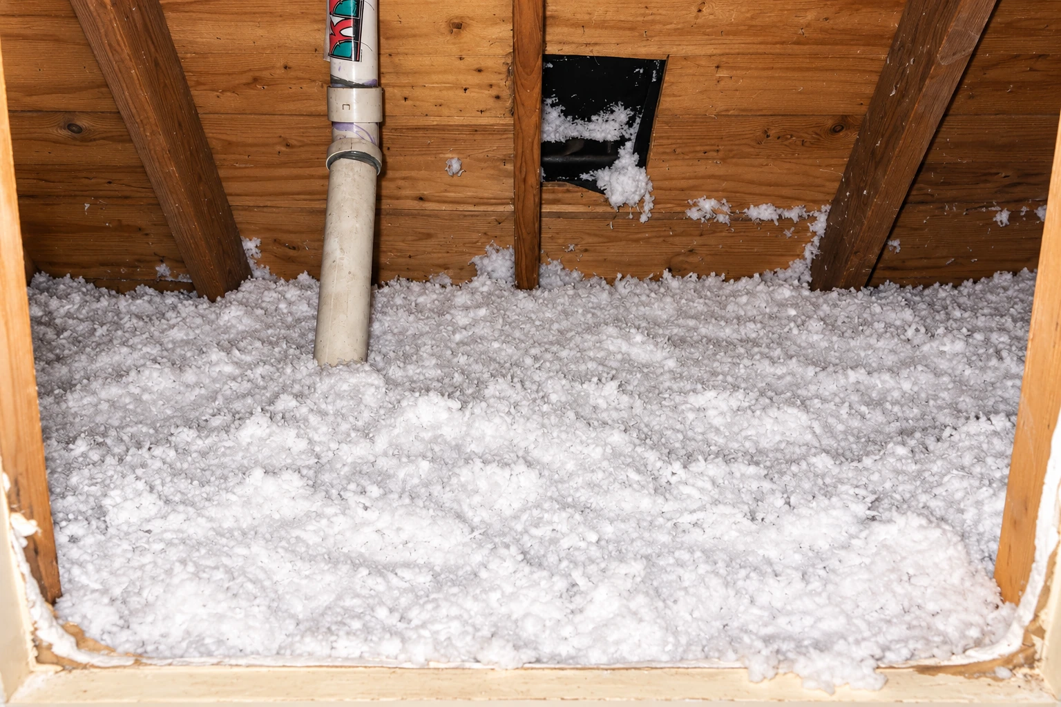 Attic floor covered with loose white blown-in insulation beneath exposed wooden rafters, with a pipe and roof opening visible.