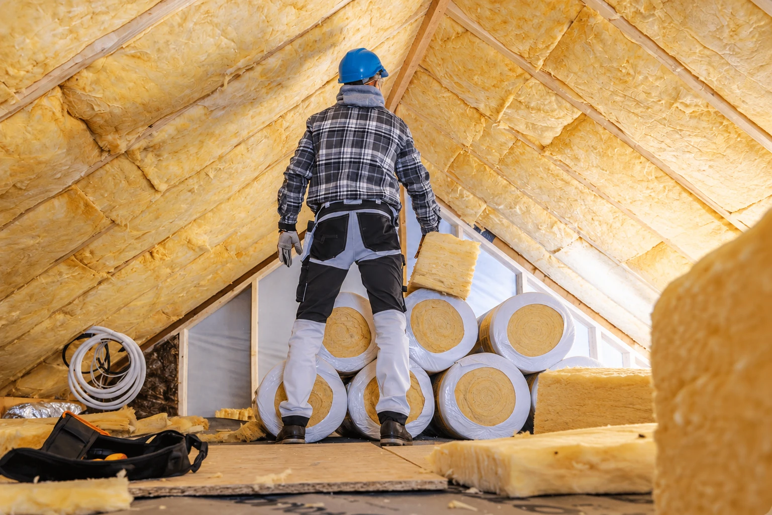 Insulation worker standing among rolls of yellow fiberglass batts in an attic with insulated roof rafters.